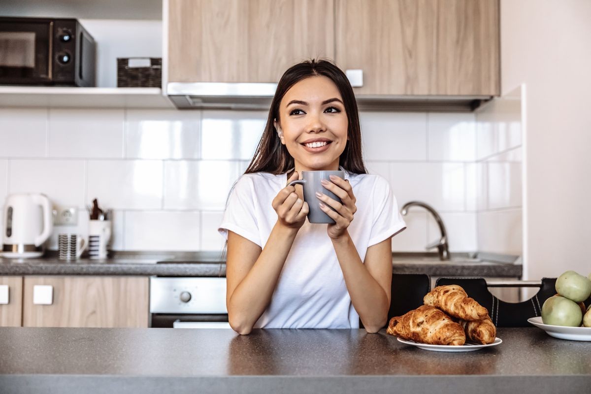  At home. Coffee time. Asian girl is holding a cup and smiling; in the kitchen